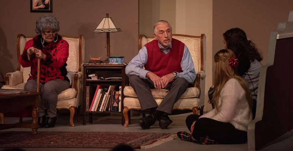 A living-room set on stage with an older couple in armchairs and two young women sitting on the floor