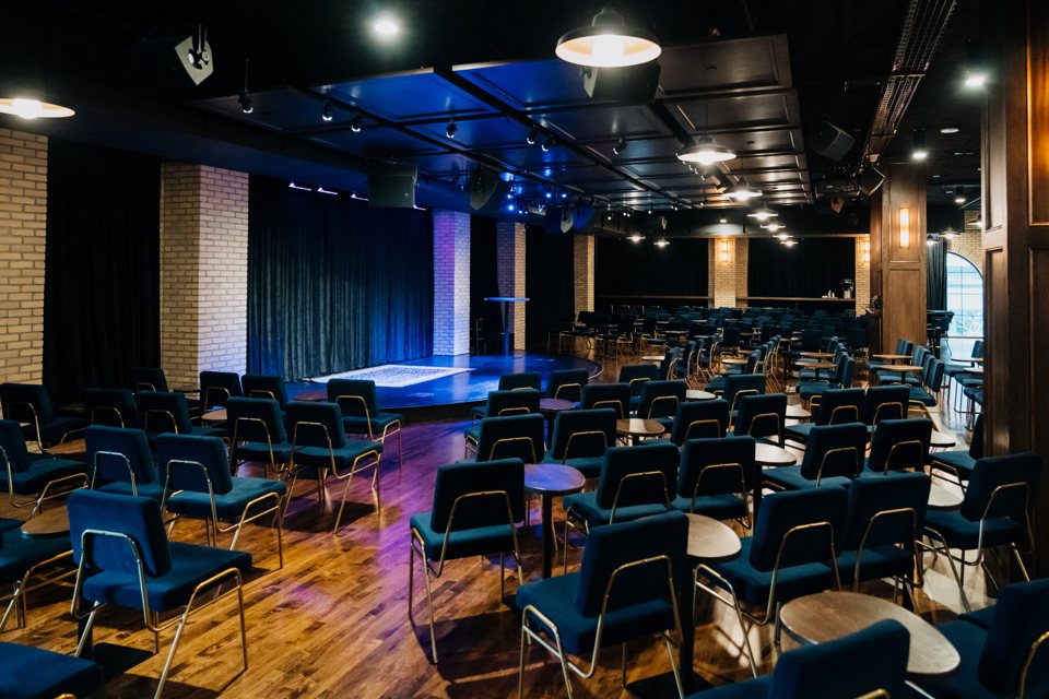 River City Improv's Midtown room with blue upholstered chairs, small tables and a stage lit in blue