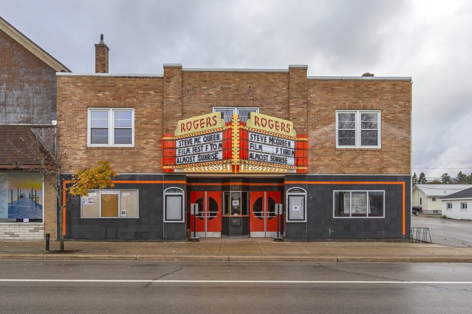 The Rogers Theater's buff-brick facade with its lit red-and-yellow ROGERS marquee and red entrance doors