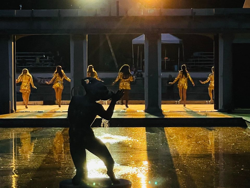 Dancers in gold sequined costumes on the outdoor stage, with a statue silhouetted in the shimmering reflecting pool in front