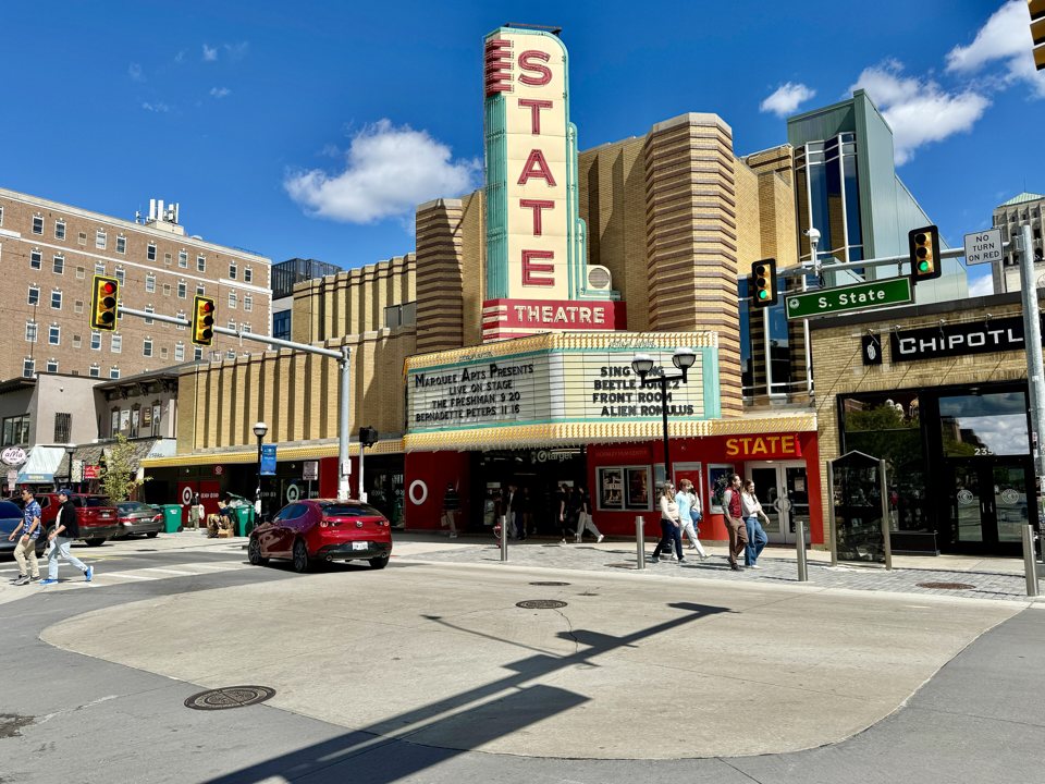 The State Theatre's vertical STATE sign and marquee on a sunny day at the corner of State Street, with people walking past