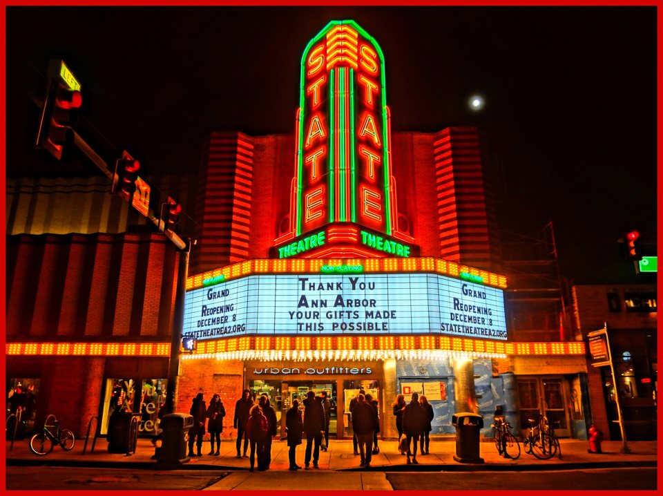 The red and green neon sign and marquee glowing at night with a thank-you message announcing the grand reopening, people on the sidewalk below