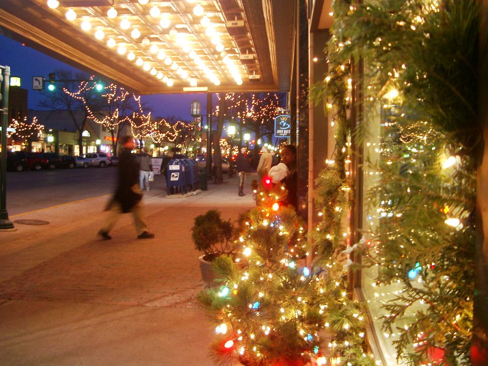 The sidewalk under the bulb-lit marquee decorated with Christmas lights, with evening passersby