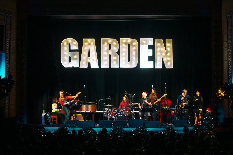 A band plays on stage beneath giant bulb-lit GARDEN letters, with holiday decorations