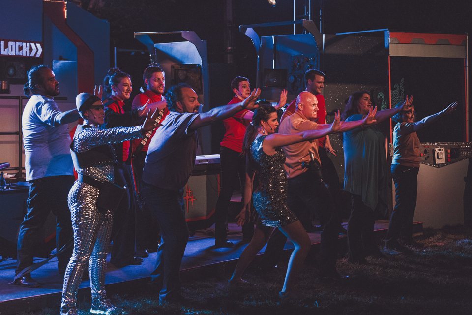 Cast members in shiny costumes dance with outstretched arms on a spaceship set on an outdoor stage