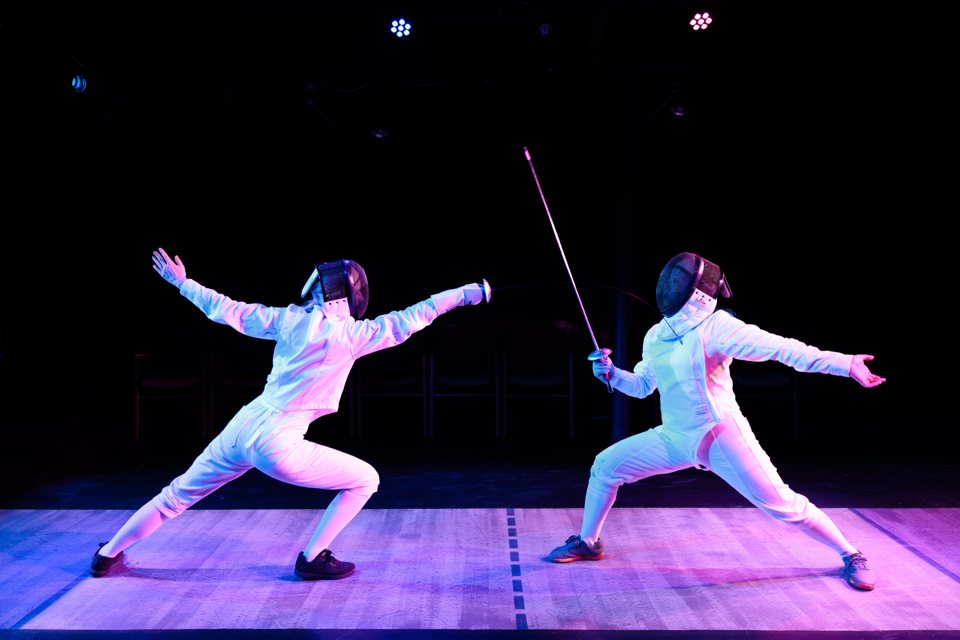 Two fencers in white face off on a stage bathed in purple light