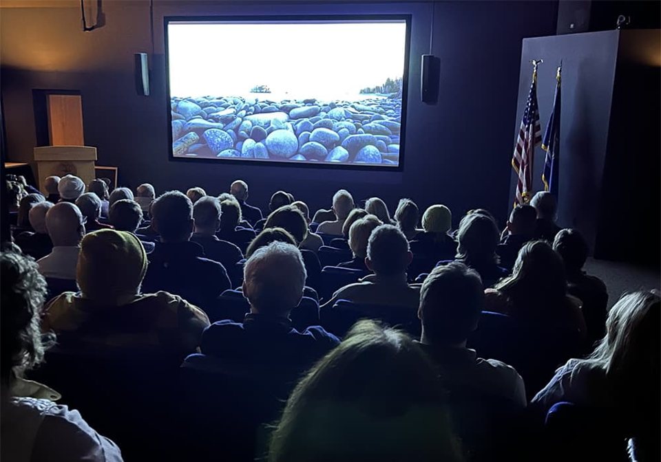 A full house watches a screening, with a rocky lakeshore on the screen