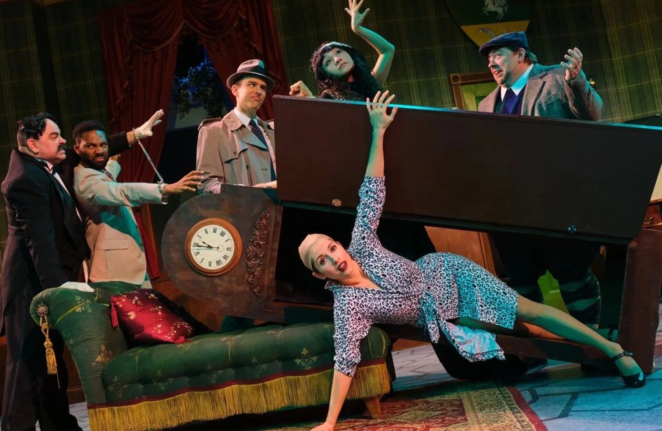 Costumed actors in hats gather around a piano while an actress in a leopard-print dress stretches out beneath it