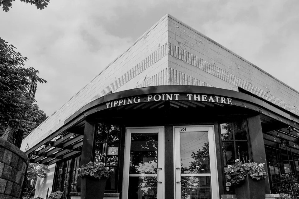 The Tipping Point Theatre's corner entrance with its curved sign and flower planters, in black and white