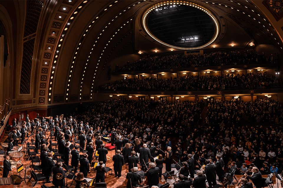 A packed Hill Auditorium as the orchestra stands for applause, under the curved light-lined ceiling and balconies