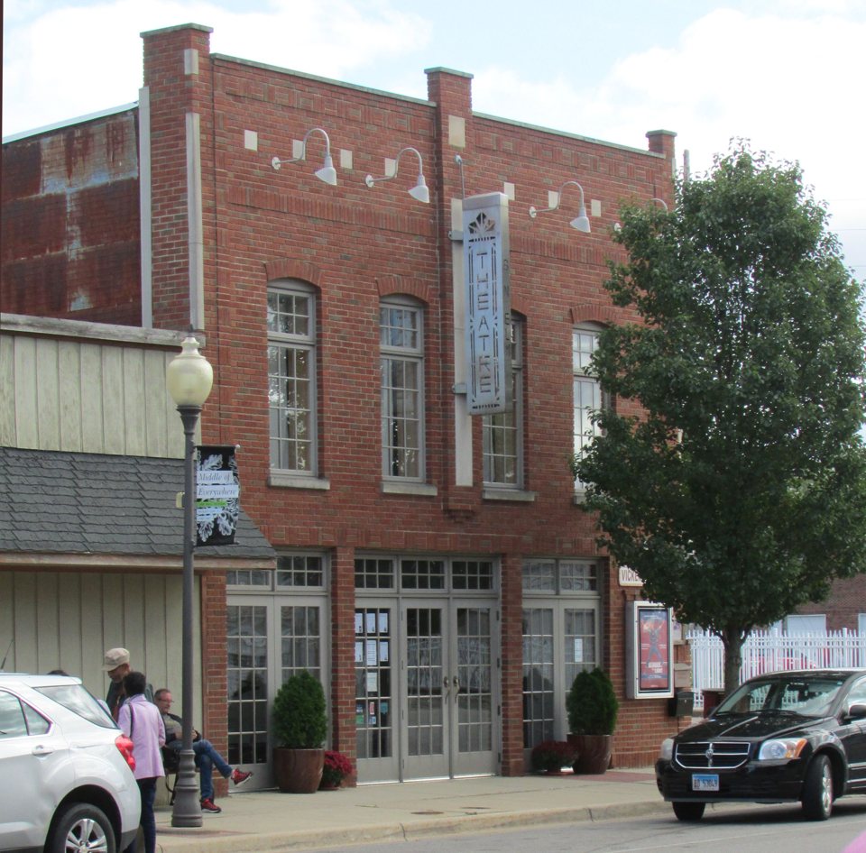 The Vickers Theatre's brick facade with tall windows and a vertical THEATRE sign, people chatting on the sidewalk