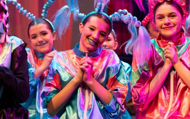 Young performers in shiny, colorful costumes and whimsical headpieces smile on stage
