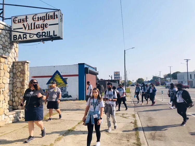 The group walks past the East English Village Bar & Grill sign on a Detroit street