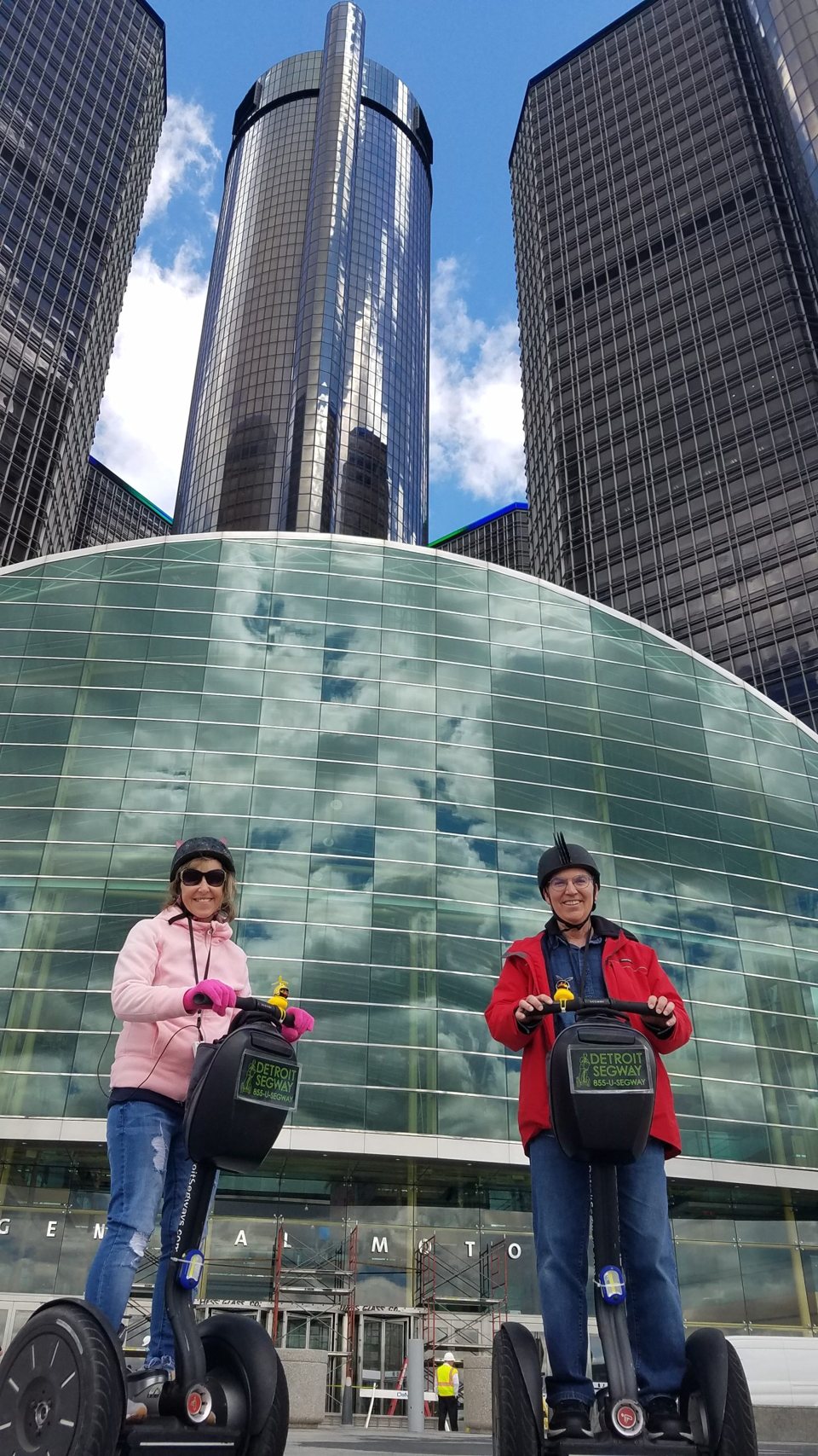 Két utas Segway-en áll a Renaissance Center tornyai előtt, a cég logójával a táskán