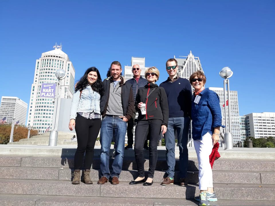 A group of six stands on the steps at Hart Plaza with downtown Detroit's skyscrapers behind them