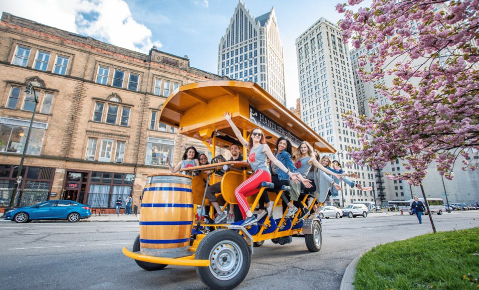 A cheerful group pedals the yellow party bike down a Detroit street, with cherry blossoms and skyscrapers behind them