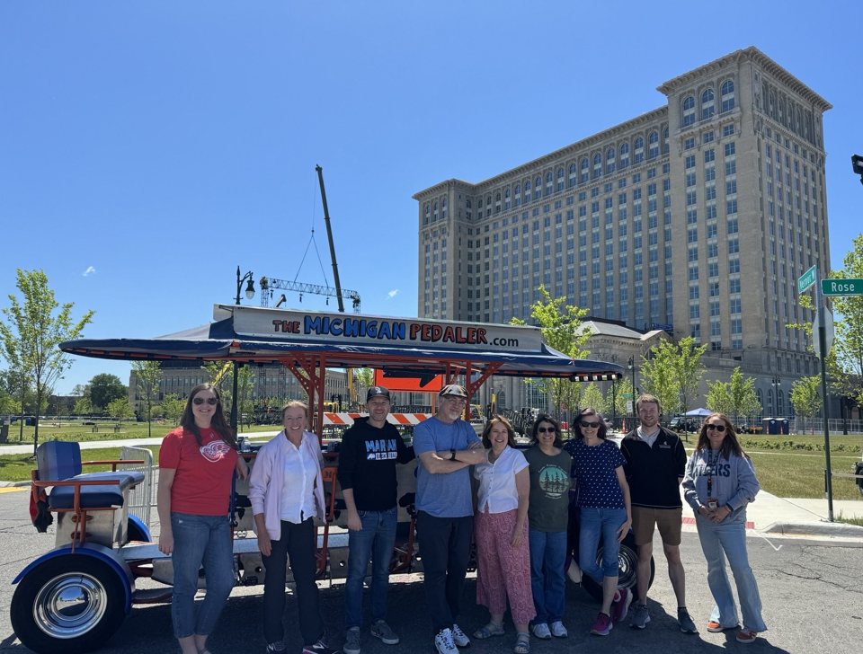 A group stands in front of the Michigan Pedaler party bike with Michigan Central Station behind them