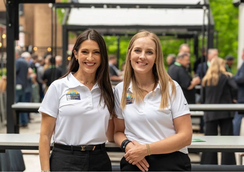 Two smiling Detroit Motor City staff members pose in branded polo shirts at an outdoor event