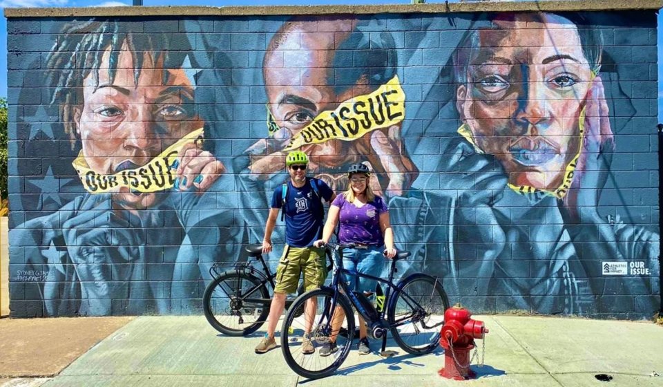 A group of cyclists pose in front of a colorful street mural in Detroit