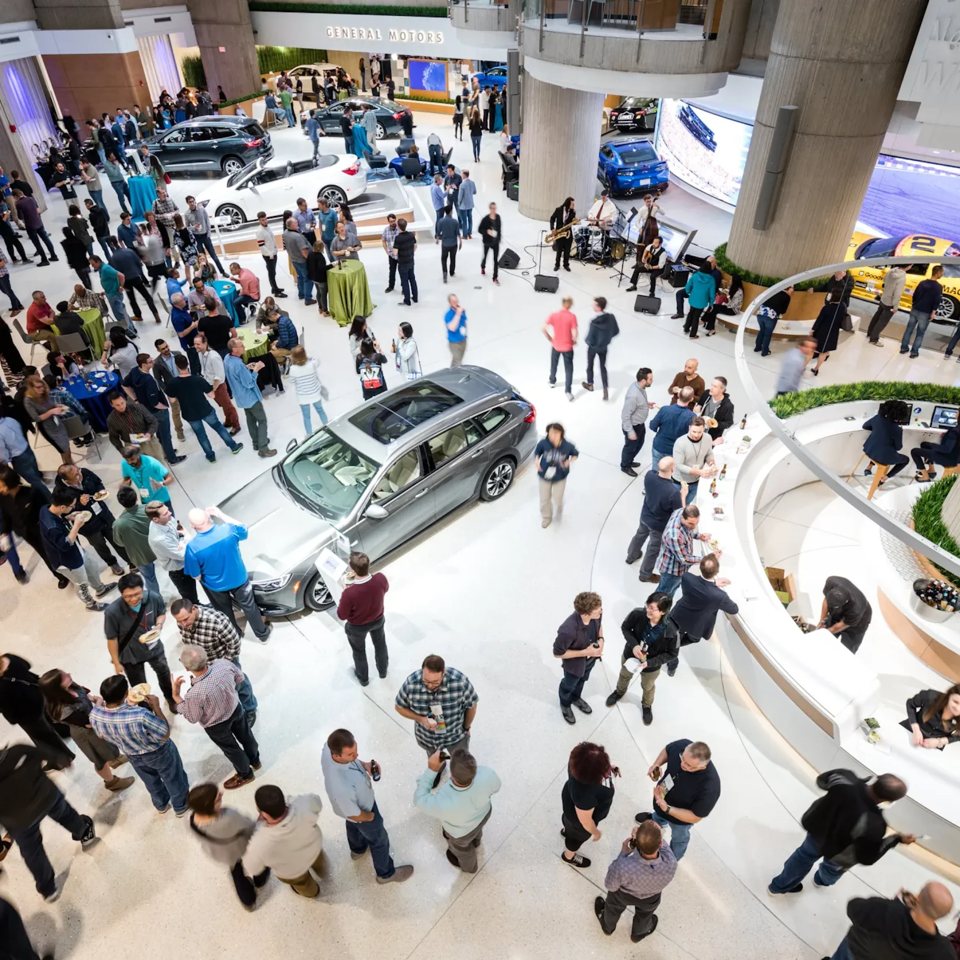 Vendégek beszélgetnek élő zenekar és kiállított autók között a General Motors épületében